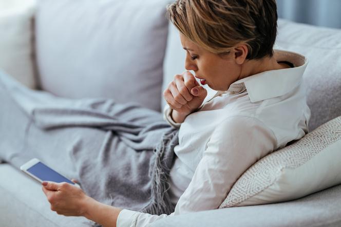 zbliżenie na kaszlącą kobietę, która leży na łóżku i korzysta z telefonu