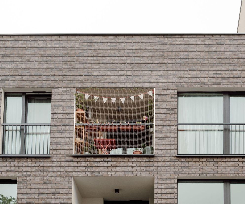 Fasada budynku kompleksu Nova Mikołowska w Katowicach, z widocznym balkonem z siatką zabezpieczającą i girlandą trójkątnych chorągiewek. Obok przeszklone okna z czarnymi ramami i balustradami. Więcej o architekturze na Architektura Murator Plus.