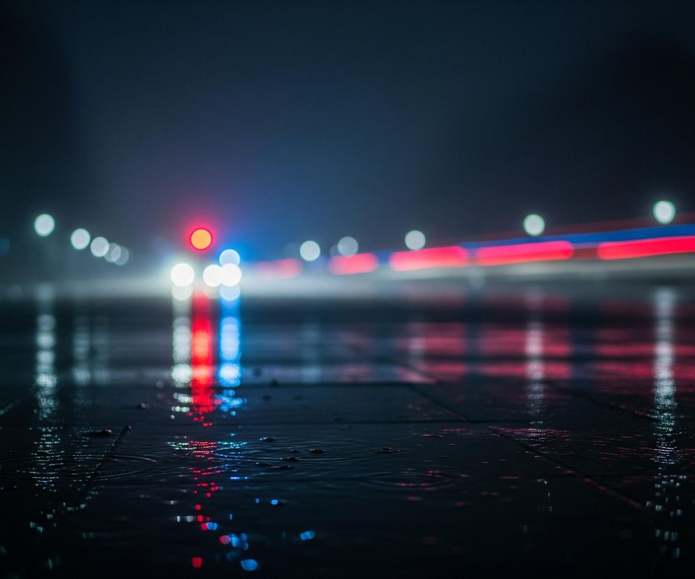 Ciemna, rozmyta scena nocna przedstawia mokrą asfaltową drogę oświetloną przez różnokolorowe światła, tworzące podłużne smugi. Na pierwszym planie widoczna jest czarna, wilgotna nawierzchnia z drobnymi kałużami i odbiciami świateł w kolorach czerwonym, niebieskim i białym. W oddali, w centrum kadru, znajdują się jaskrawe, rozmazane światła, w tym wyraźne czerwone koło i niebieskie pasmo, sugerujące źródło światła pojazdu, otoczone mniejszymi, białymi punktami. W prawym górnym rogu obrazu, nad drogą, widać poziome smugi światła w kolorach czerwonym i niebieskim, odbijające się częściowo na mokrej powierzchni.