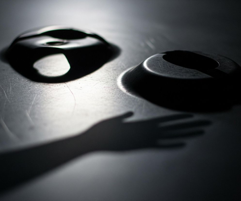 Dwie ciemne, owalne plamy o nieregularnych krawędziach, leżące na jasnoszarej, lekko porysowanej powierzchni. Górna plama ma wewnątrz wyraźne wgłębienie, a obie otoczone są jasnymi refleksami światła, które podkreślają ich kształt. W lewym dolnym rogu zdjęcia rozciąga się długi, ciemny cień ludzkiej dłoni z rozpostartymi palcami. Całość utrzymana jest w ciemnych, monochromatycznych barwach, z ostrym kontrastem między jasnymi refleksami a głębokimi cieniami.