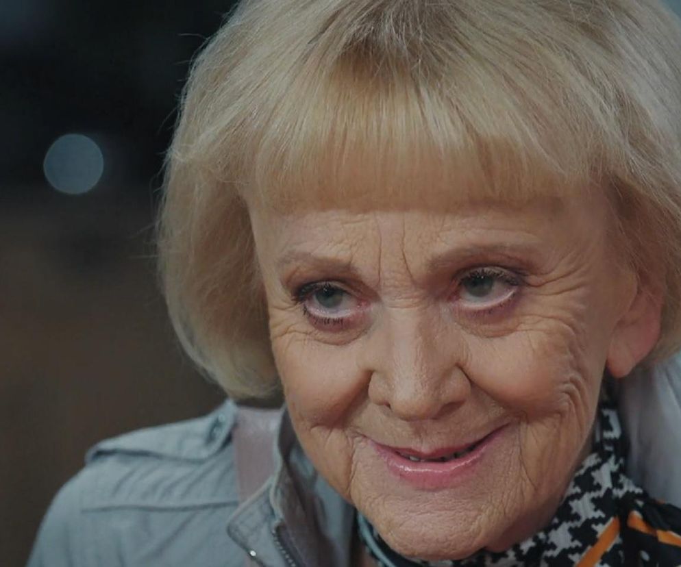 Blondwłosa Małgorzata Rożniatowska jako Kisielowa z serialu M jak miłość z delikatnym uśmiechem, ubrana w jasnoszarą kurtkę i wzorzystą apaszkę, prezentuje determinację w odcinku 1878. Więcej o losach bohaterów przeczytasz na Super Seriale.