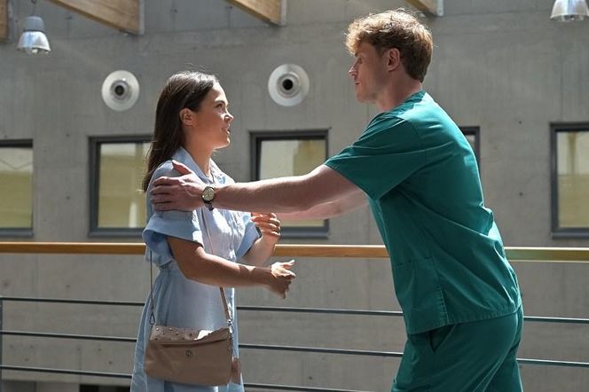 Radek (Karol Biskup) w zielonym stroju medycznym dotyka ramienia Julity (Dagmara Bryzek) w jasnoniebieskiej sukience. Stoją na tle nowoczesnego budynku, z oknami i betonową ścianą. Scena odzwierciedla napięcie i oddalenie między bohaterami serialu Pierwsza miłość, o którym więcej przeczytasz na Super Seriale.