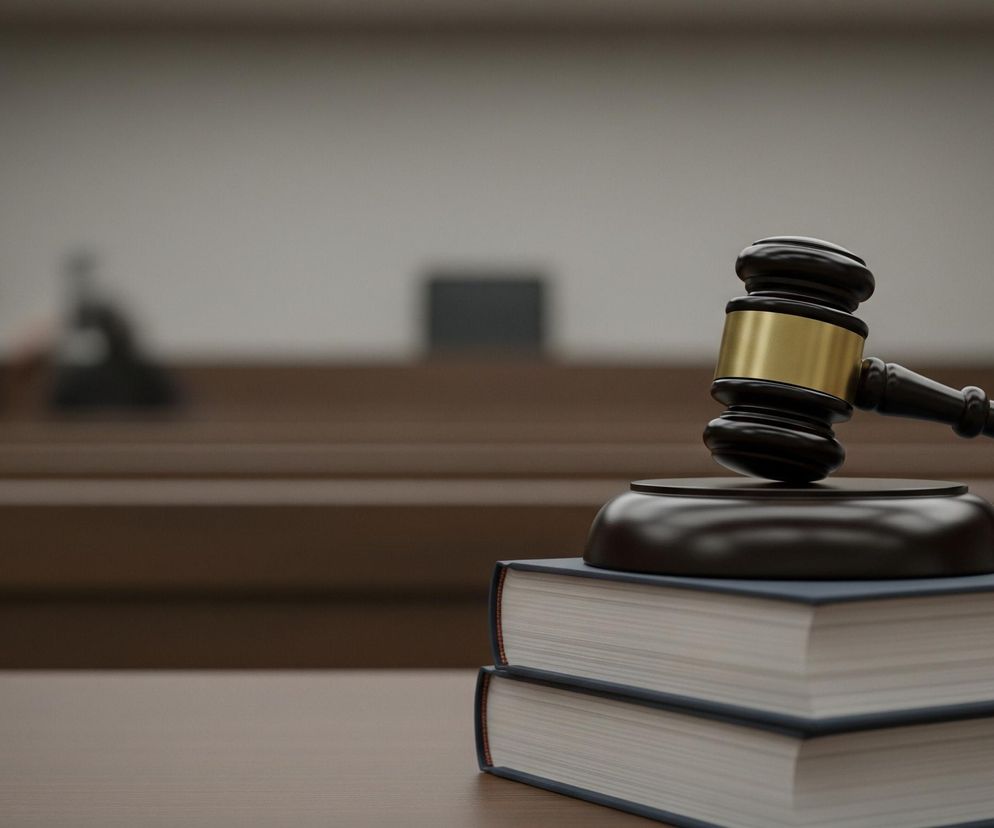 Na pierwszym planie, z prawej strony, leży czarny drewniany młotek sędziowski ze złotym pasem na głowicy, oparty na okrągłej podstawce. Pod podstawką znajdują się dwa ułożone jeden na drugim tomy książek w granatowych oprawach z białymi kartkami, spoczywające na brązowym, drewnianym stole. Tło jest rozmyte, ukazując salę sądową z rzędami drewnianych ław i jasnoszarymi ścianami.