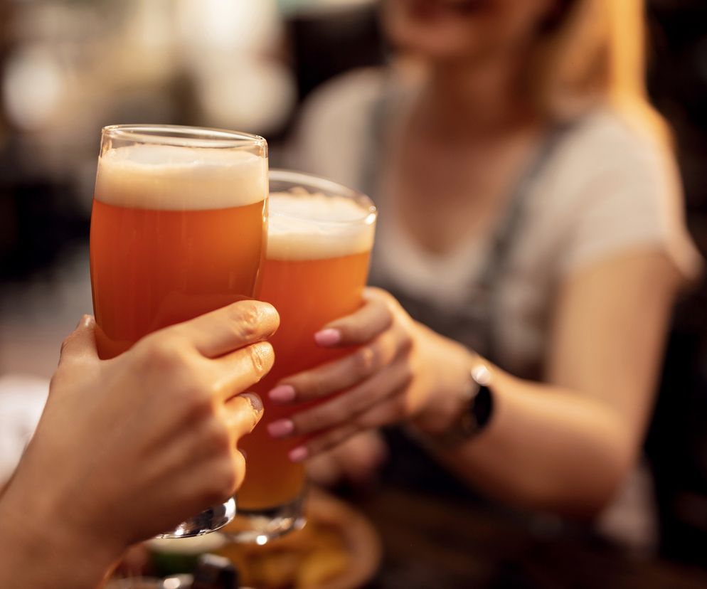 Dwie dłonie, jedna z różowymi paznokciami, wznoszą szklanki jasnego piwa z pianą w geście toastu, na rozmytym tle uśmiechniętej osoby. Scena symbolizuje społeczne konteksty picia alkoholu, o których możesz przeczytać na portalu Poradnik Zdrowie.