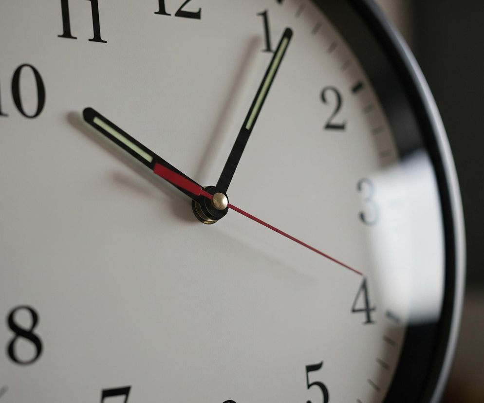 Duży, okrągły zegar ścienny z czarną ramką dominuje centralną część obrazu. Jego biała tarcza jest wyraźnie widoczna, ze wszystkimi czarnymi cyframi od 1 do 12 oraz podziałkami minutowymi. Na tarczy znajdują się trzy wskazówki: czarna wskazówka godzinowa z fluorescencyjnym paskiem skierowana na cyfrę 10, krótsza czarna wskazówka minutowa również z fluorescencyjnym paskiem skierowana na cyfrę 1, oraz cienka, czerwona sekundowa wskazówka wskazująca na około 4. W centrum tarczy, gdzie wszystkie wskazówki się łączą, widoczny jest mały, złoty element. Tło za zegarem jest nieostre, ukazując rozmyte kształty jasnego okna po prawej stronie i ciemniejszej ściany po lewej.