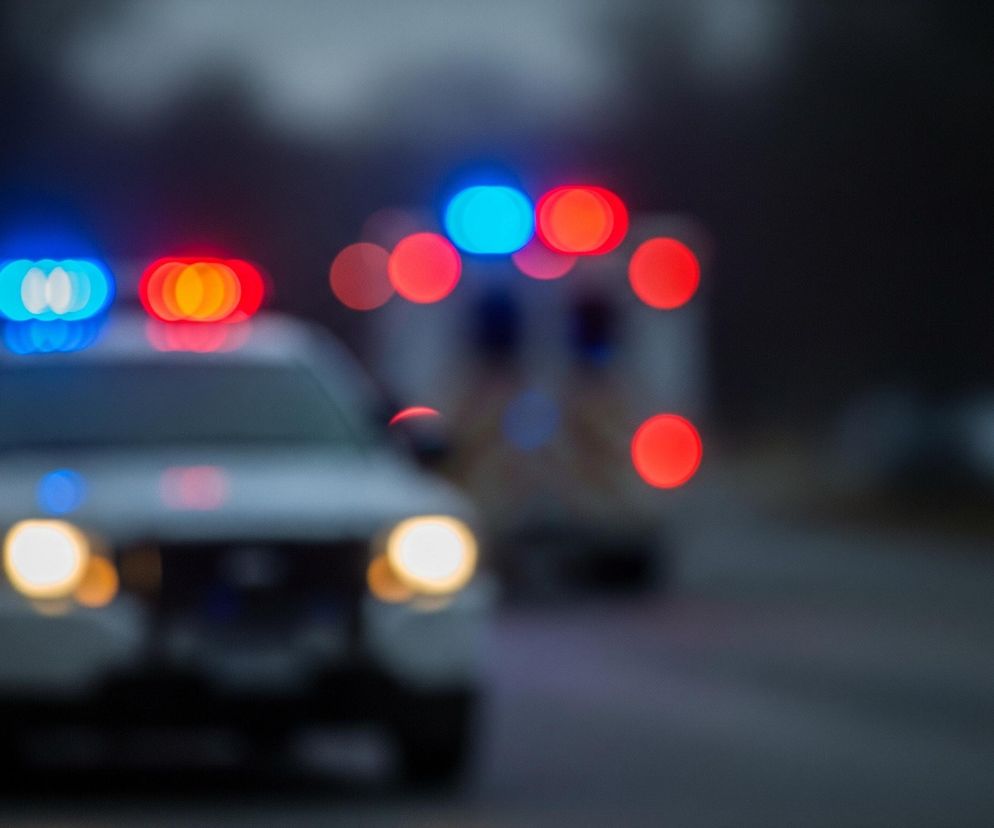 Zamazane ujęcie dwóch pojazdów ratunkowych jadących po drodze w tle zmierzchu. Na pierwszym planie znajduje się biały samochód policyjny z włączonymi, rozmazanymi światłami awaryjnymi w kolorze niebieskim i czerwonym na dachu oraz włączonymi przednimi reflektorami. Za nim, również w rozmazanym ujęciu, jedzie karetka pogotowia, której górna część dachu oraz tył są białe z widocznymi czerwonymi i niebieskimi światłami sygnalizacyjnymi. Droga jest ciemnoszara, a tło stanowią rozmazane, ciemne sylwetki drzew lub innych obiektów.