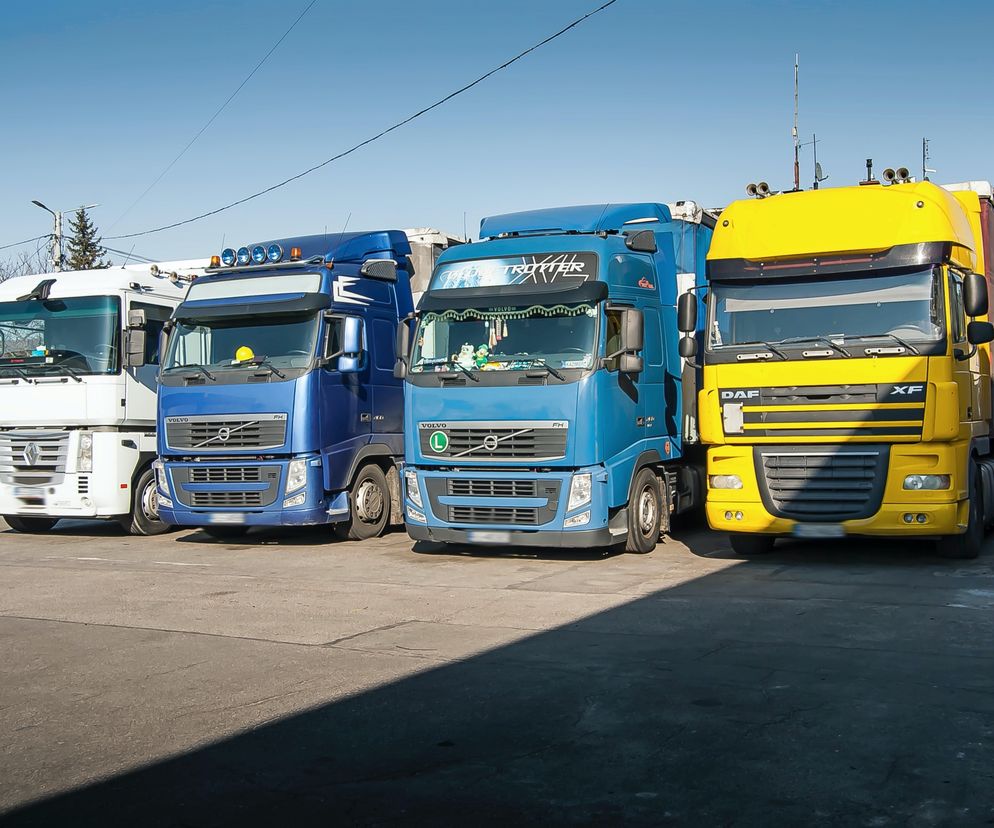 Rząd ciężarówek (białej, dwóch niebieskich i żółtej) na parkingu, obrazująca rosnące zadłużenie w transporcie drogowym. Problemy branży TSL, w tym spadek przewozów i zatory płatnicze, znajdziesz na Super Biznes.