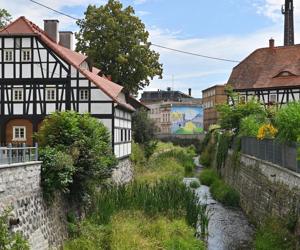 Malowniczy dom przysłupowy w Bogatyni z czerwonym dachem i biało-czarnym szachulcem nad zielenią przy wąskim strumieniu. W tle drugi podobny budynek. O zabytkowej architekturze Bogatyni, w tym domach przysłupowych, przeczytasz na portalu Architektura Murator Plus.