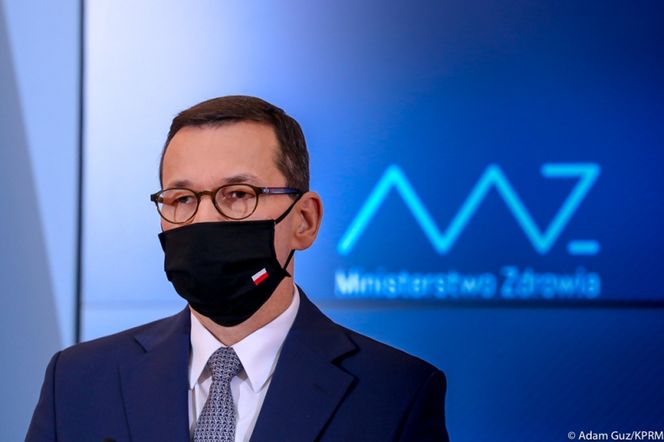 Mateusz Morawiecki w masce ochronnej z biało-czerwoną flagą na tle niebieskiego ekranu z logo Ministerstwa Zdrowia. Premier opowiada o Narodowym Programie Szczepień, jego szczegółach i bezpieczeństwie, o czym więcej przeczytasz na Poradnik Zdrowie.