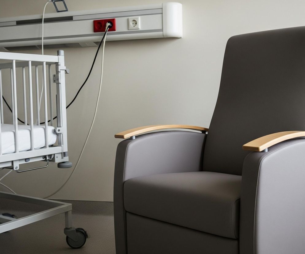 Na pierwszym planie po prawej stronie znajduje się duże, szare, tapicerowane krzesło z drewnianymi podłokietnikami, skierowane częściowo w lewo. Po lewej stronie widać białe, metalowe łóżeczko dziecięce z materacem, wyposażone w kółka. Na ścianie za meblami, na wysokości wzroku, zamontowana jest pozioma listwa ścienna z gniazdkami elektrycznymi i innymi przyłączami, w tym jednym czerwonym. Na dalszym planie po prawej stronie stoi biały grzejnik żeberkowy.