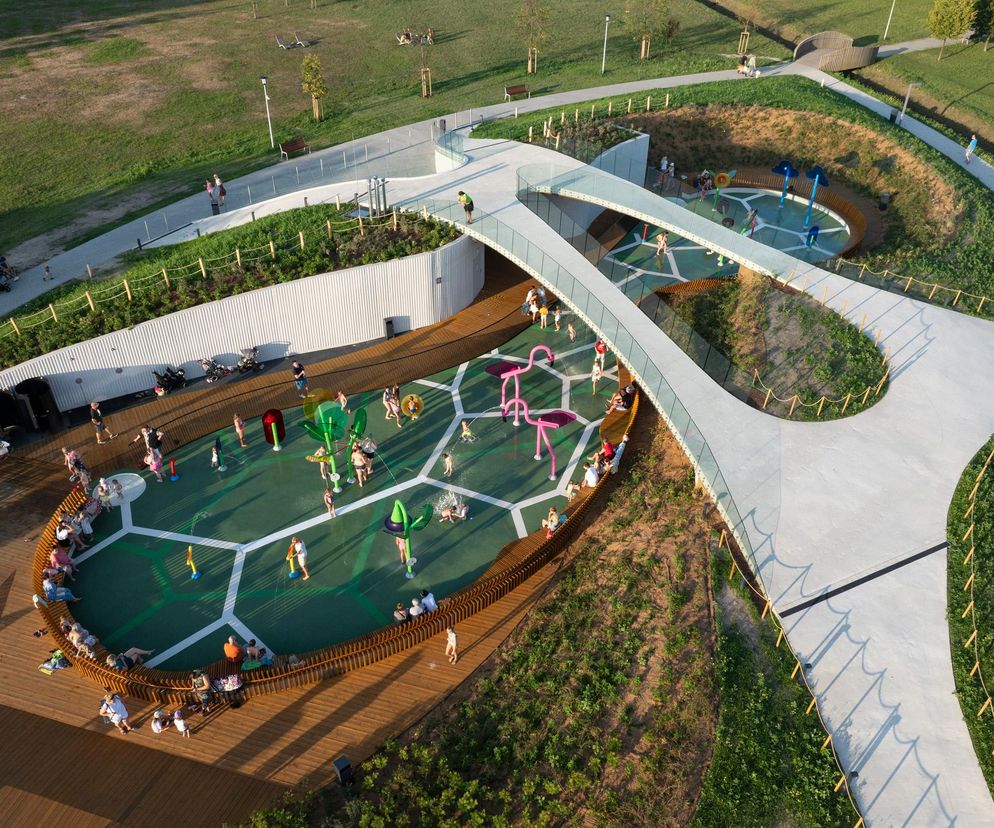 Ujęcie z drona przedstawiające wodny plac zabaw w Parku Jaworek w Tychach z aktywnymi dziećmi i dorosłymi. Widoczne są mostki, drewniane siedziska oraz zielone tereny rekreacyjne. Więcej o tej realizacji znajdziesz na Architektura Murator Plus.