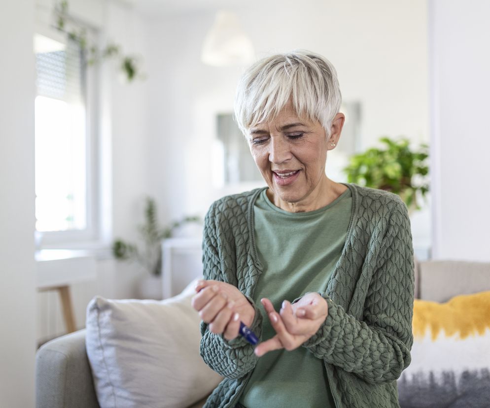 Starsza kobieta z krótkimi, siwymi włosami i uśmiechem na twarzy, mierząca poziom cukru we krwi glukometrem w domowym zaciszu, siedząc na kanapie. Artykuły na temat monitorowania zdrowia, w tym poziomu cukru, można znaleźć na portalu Poradnik Zdrowie.