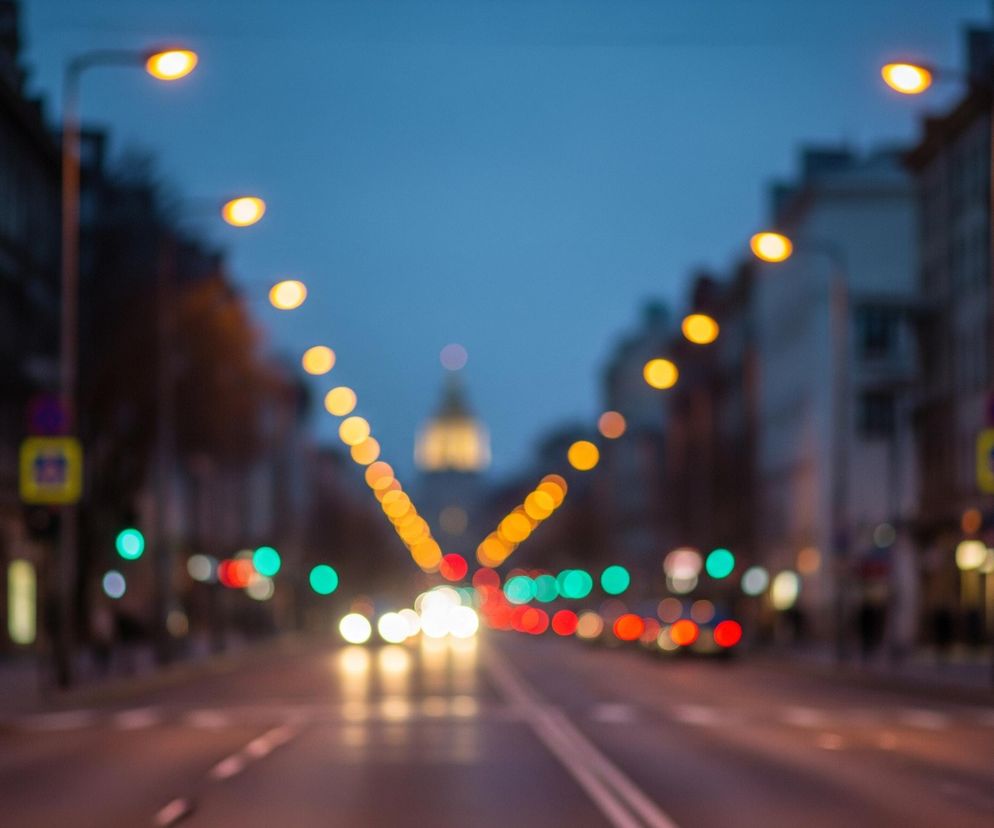 Centralnie na obrazie dominuje rozmyta perspektywa ulicy miejskiej o zmierzchu lub w nocy, z rzędami latarni po obu stronach. Światła latarni ulicznych są widoczne jako owalne, pomarańczowo-żółte kręgi bokeh, ciągnące się w głąb obrazu. Na końcu ulicy, na horyzoncie, widać rozmyty zarys budowli z podświetloną kopułą.

Ulica jest oświetlona przez reflektory nadjeżdżających samochodów, które tworzą jasne, białe plamy bokeh w centrum obrazu, oraz przez tylne światła pojazdów, widoczne jako czerwone kręgi. Zielone światła sygnalizacji świetlnej również są widoczne po obu stronach drogi, jako wyraźne, zielone plamy. Po lewej i prawej stronie ulicy widać zarysy budynków mieszkalnych, ich okna i fasady są rozmyte i słabo widoczne w ciemności.