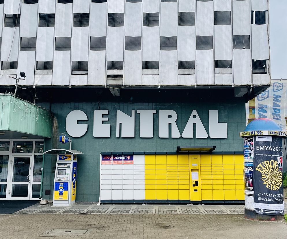 Front domu handlowego Central w Białymstoku, ukazujący charakterystyczną ażurową elewację z aluminiowych łusek oraz logo CENTRAL na zielonej ścianie. Przed wejściem widoczne paczkomaty, bankomat i niebieski SUV. O modernizacji budynku przeczytasz na Architektura Murator Plus.