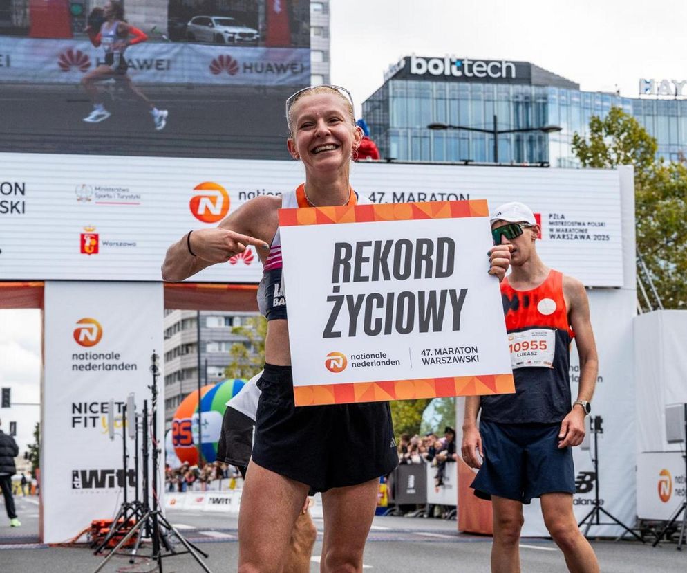 Szczęśliwa biegaczka z tabliczką Rekord Życiowy po ukończeniu 47. Nationale-Nederlanden Maratonu Warszawskiego, obok niej inny uczestnik, w tle tablice reklamowe i duży ekran wyświetlający wyniki. Więcej o sukcesie wydarzenia przeczytasz na Super Biznes.