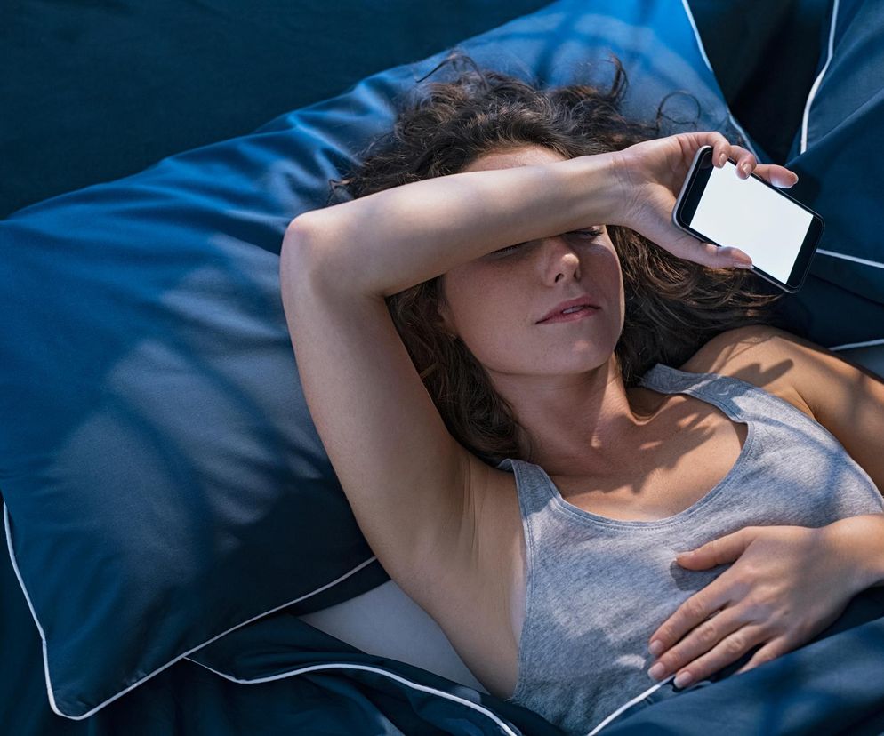 Kobieta leży w łóżku pod ciemnoniebieską pościelą z białą lamówką, trzymając telefon z białym ekranem nad czołem, zasłaniając oczy dłonią. Ta scena symbolizuje problemy ze snem i używanie telefonu przed zaśnięciem, o czym można przeczytać na portalu Poradnik Zdrowie.