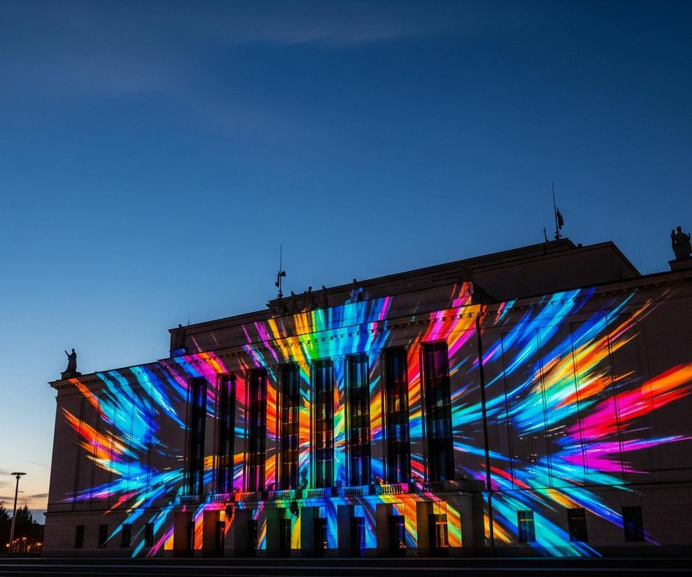 Duży budynek o klasycznej architekturze, z kolumnami i posągami na dachu, jest podświetlony wielobarwną projekcją. Kolory, takie jak niebieski, zielony, żółty, pomarańczowy, czerwony i fioletowy, tworzą dynamiczne, rozchodzące się smugi i paski, rozświetlające fasadę. Scena przedstawia się o zmierzchu, z ciemniejącym niebieskim niebem, na którym widać jeden jasny punkt, prawdopodobnie gwiazdę lub planetę. W dolnej części obrazu widoczne są schody prowadzące do budynku oraz fragmenty oświetlenia ulicznego.