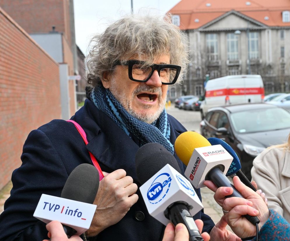 Janusz Palikot z siwymi, kręconymi włosami i w dużych okularach, w granatowym płaszczu i błękitnym szaliku, przemawia przed mikrofonami stacji TVN24, TVP Info i Radia Wrocław. Z boku widać fragment kobiety. Więcej informacji na Super Biznes.