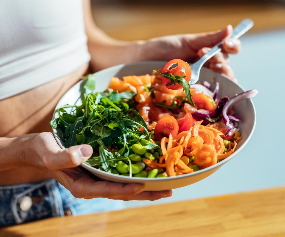 Kobieta w białym topie i dżinsach trzyma miskę pełną zdrowych składników: rukoli, pomidorków koktajlowych, edamame, startej marchewki i czerwonej cebuli. Na widelcu kawałek pomidora z rukolą. Zdjęcie symbolizuje zdrową dietę i wpływ leków na odczuwanie smaków, o czym przeczytasz na Poradnik Zdrowie.
