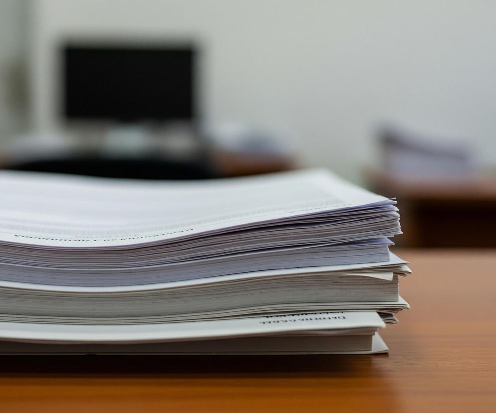 Na pierwszym planie, z lewej strony, znajduje się wysoki stos białych dokumentów lub kartek papieru z delikatnymi nadrukami tekstowymi. Stos leży na brązowym, drewnianym biurku, którego faktura jest widoczna. Tło jest rozmyte, ukazując jaśniejszą ścianę i ciemne, niewyraźne kształty, sugerujące obecność monitora komputera oraz innych obiektów biurowych.