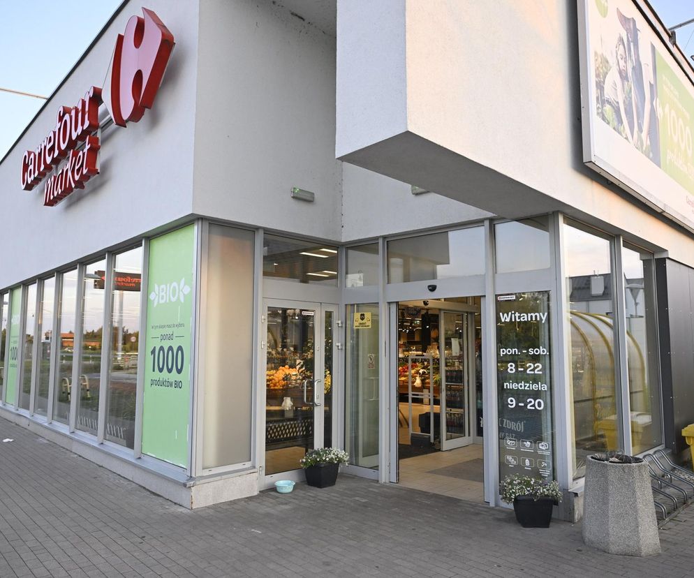 Wejście do marketu Carrefour z dużym logo i przeszklonymi drzwiami, obok których widnieją godziny otwarcia wskazujące na handel również w niedziele. Widoczne plakaty z ofertą produktów BIO. Artykuł na Super Biznes informuje o zmianach w handlu w niedziele.