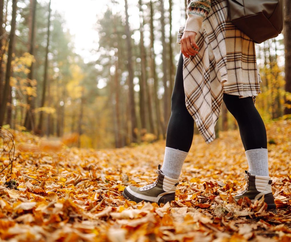 Kobieta w sportowym obuwiu, czarnych legginsach i koszuli w kratę spaceruje po lesie, pokrytym warstwą pomarańczowych liści. W tle widać pnie drzew. To symboliczna grafika dotycząca korzyści ciągłego spaceru dla serca, o czym przeczytasz na Poradniku Zdrowie.