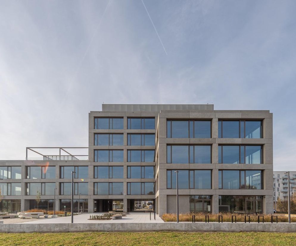 Nowoczesny biurowiec na Nowych Żernikach we Wrocławiu, projektu Maćków Pracownia Projektowa, z elewacją z szarego betonu i dużymi oknami. Budynek prezentuje się elegancko i funkcjonalnie. Więcej o takich obiektach przeczytasz na Architektura Murator Plus.