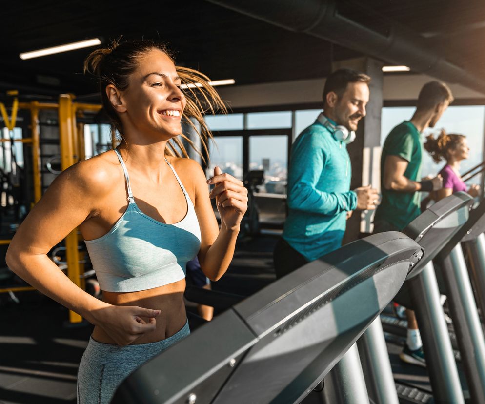 zbliżenie na uśmiechniętą kobietę, która biegnie na bieżni podczas treningu na siłowni