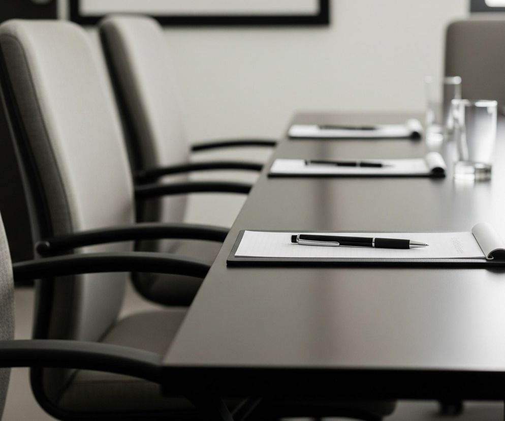 Rząd krzeseł z szarymi, tekstylnymi oparciami i czarnymi podłokietnikami rozciąga się w lewo, z ostrością na najbliższym krześle. Po prawej stronie widać ciemny, prostokątny stół, na którym leżą podkładki z białym papierem i długopisami, a także szklanki, niektóre z niewielką ilością wody. Tło jest jasne, z niewyraźnym zarysem ciemnej ramy obrazu lub okna w górnej części.