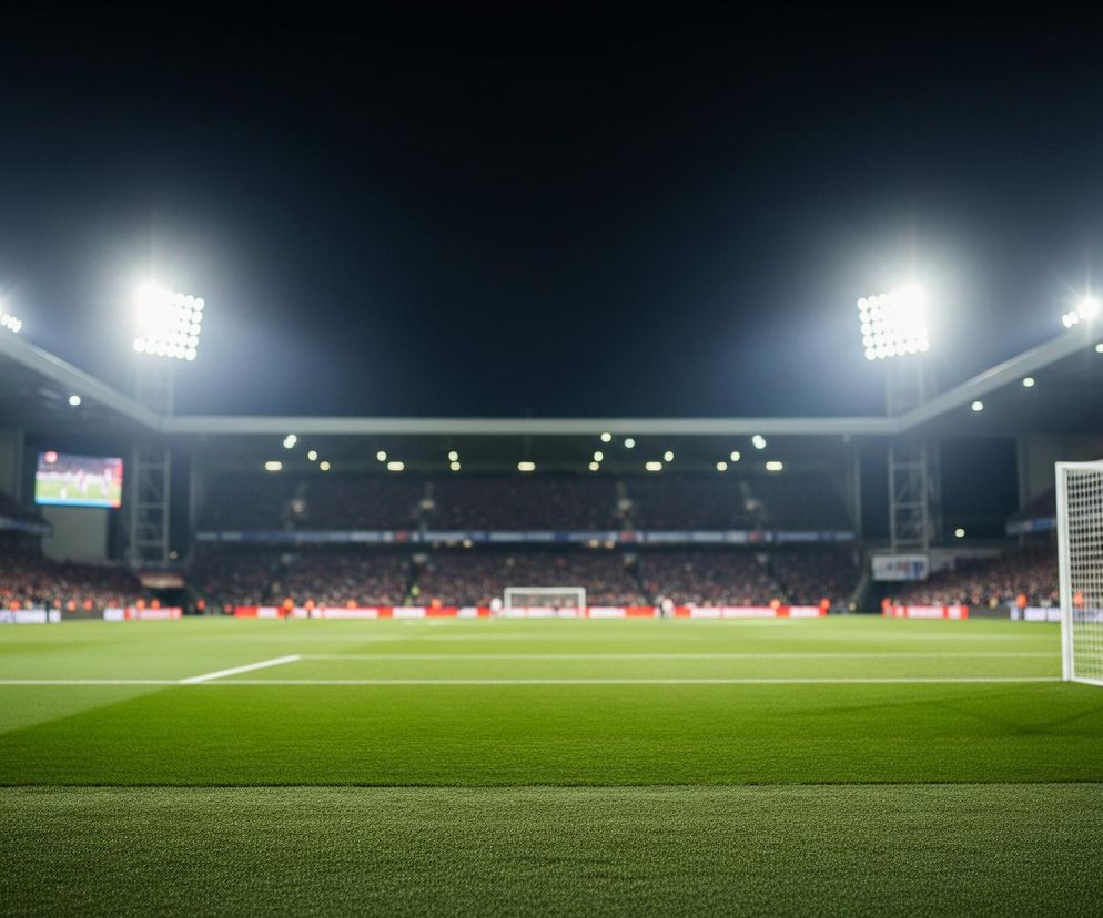 Głębokie zielone pole stadionu z jasnymi białymi liniami dominuje na pierwszym planie, rozciągając się w kierunku odległego, niewyraźnego tłumu. Na prawym planie stoi biała siatka bramkowa z czarnymi słupkami. W tle, na trybunach, widać zgromadzonych ludzi, a po lewej stronie duży ekran wyświetla dynamiczne ujęcie. Górna część obrazu to ciemne, nocne niebo, rozświetlone licznymi, intensywnie świecącymi reflektorami stadionu, tworzącymi jasne blaski światła.