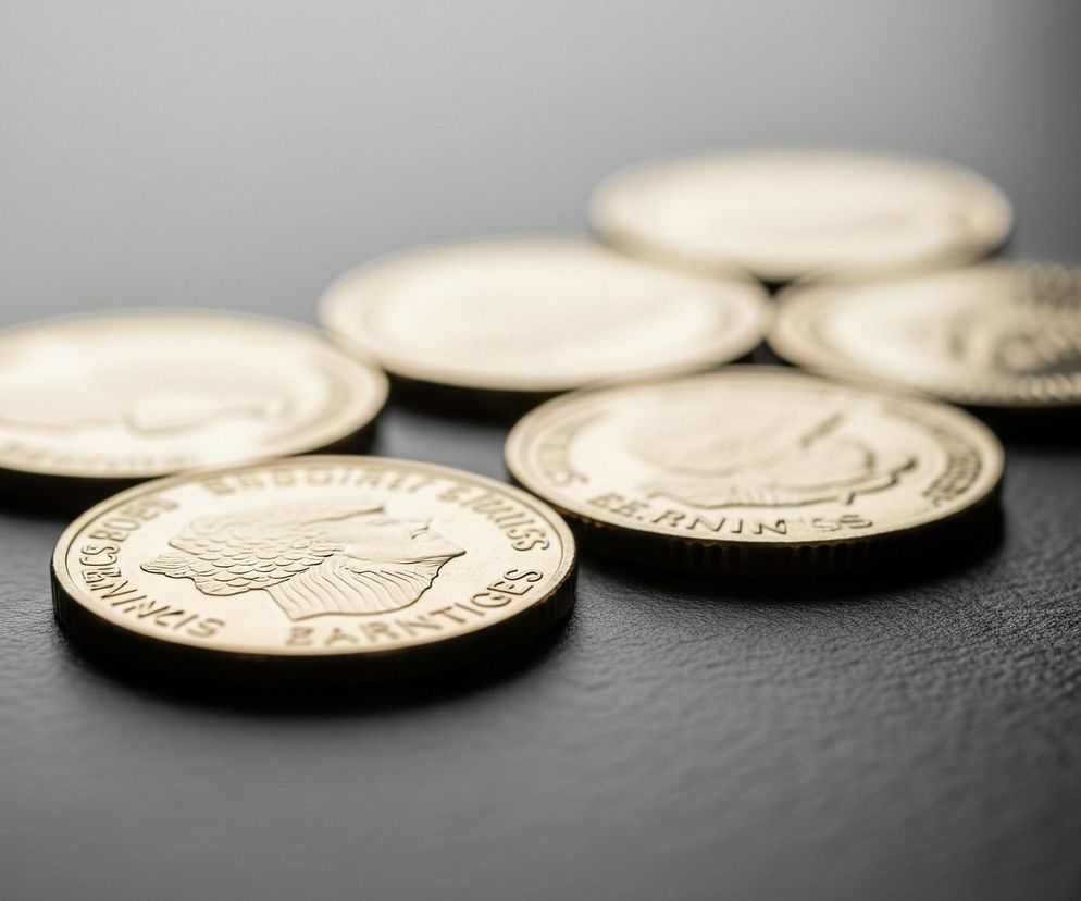 Na ciemnej, szorstkiej powierzchni leży sześć okrągłych, metalicznych monet w złotym kolorze. Jedna z monet, znajdująca się najbliżej obiektywu, jest wyraźnie ostra i przedstawia profil głowy oraz tekst wokół niej. Pozostałe monety są rozmyte, ułożone w formację przypominającą trójkąt lub kwiat, z trzema widocznymi w górnej części kadru i dwoma w środkowej części, częściowo zasłoniętymi przez monetę na pierwszym planie. Światło pada z lewej strony, tworząc delikatne refleksy na powierzchni monet i cienie na tle.