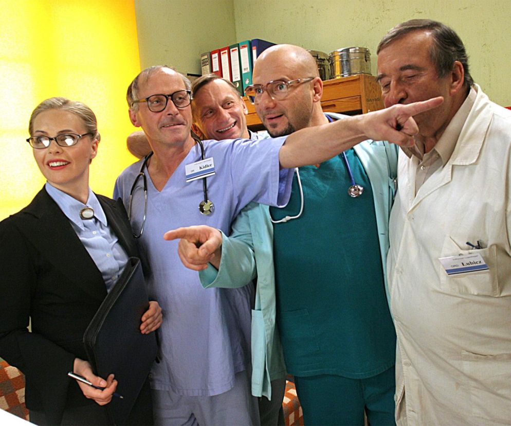 Grupowe zdjęcie aktorów z serialu Daleko od noszy – Hanny Śleszyńskiej w czarnym żakiecie, Pawła Wawrzeckiego w niebieskim uniformie chirurga z identyfikatorem Kidler, Piotra Gąsowskiego w zielonym stroju medycznym oraz Krzysztofa Kowalewskiego w białym fartuchu z plakietką Lubicz. Artykuł o kulisach produkcji serialu znajdziesz na Super Seriale.