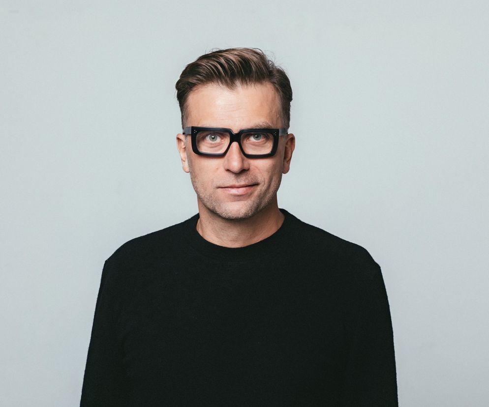 Portret Tomka Rygalika, wybitnego projektanta i edukatora, z krótkimi, zaczesanymi włosami i charakterystycznymi, grubymi, czarnymi okularami, ubranego w czarny sweter, na neutralnym jasnym tle. Więcej o jego działalności przeczytasz na Architektura Murator Plus.