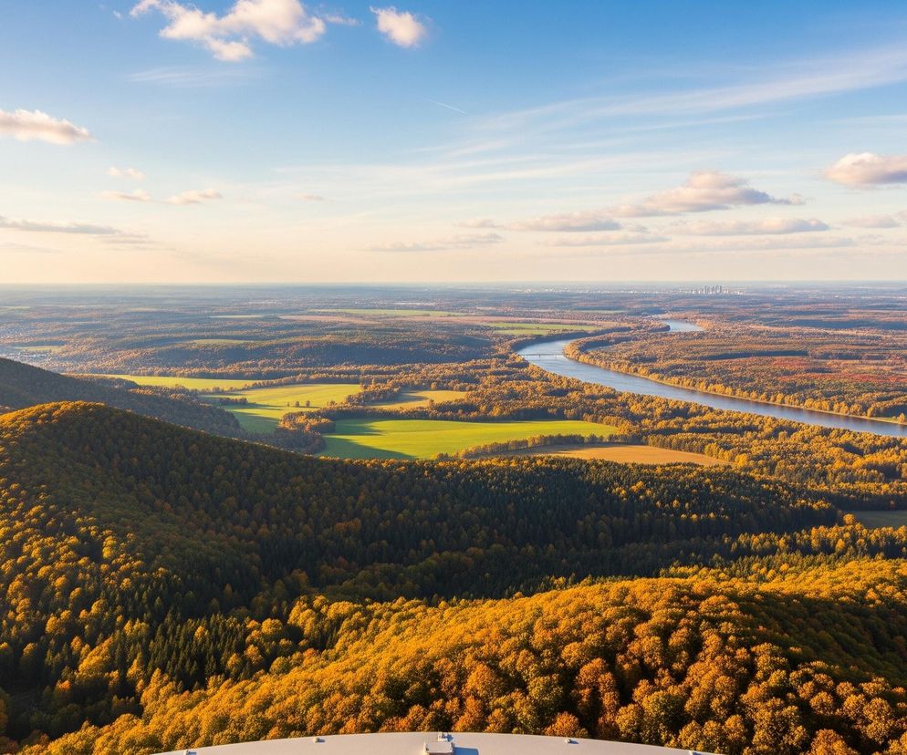 Rozległy krajobraz widziany z lotu ptaka rozciąga się pod błękitnym niebem z rozproszonymi białymi chmurami. Na pierwszym planie dominują zalesione wzgórza, pokryte gęstymi drzewami o jesiennych, złocisto-pomarańczowych liściach, z ciemniejszymi partiami iglastymi w zagłębieniach. W oddali wijąca się rzeka odbija niebieskawe światło, otoczona rozległymi polami uprawnymi w odcieniach zieleni i beżu, oraz dalszymi pasmami lasów. Na horyzoncie, po prawej stronie, majaczy zarys miejskiej zabudowy.