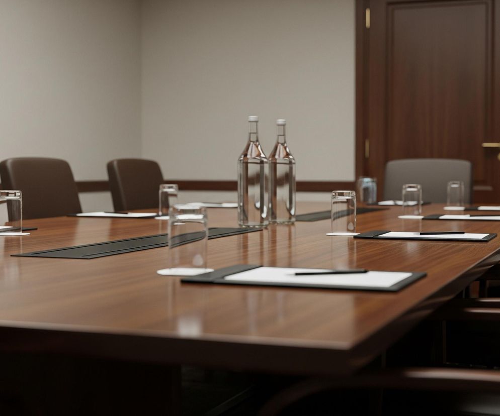 Duży, lśniący, brązowy stół konferencyjny dominuje w centrum kadru, rozciągając się od lewej krawędzi ku prawemu dolnemu rogowi. Na stole rozmieszczone są szklanki i butelki wody, a także podkładki z białym papierem i czarnymi długopisami, sugerujące gotowość do spotkania. Wokół stołu ustawione są skórzane krzesła w odcieniach brązu, a w tle widoczne są jasnoszare ściany, drewniane drzwi i rama obrazu.