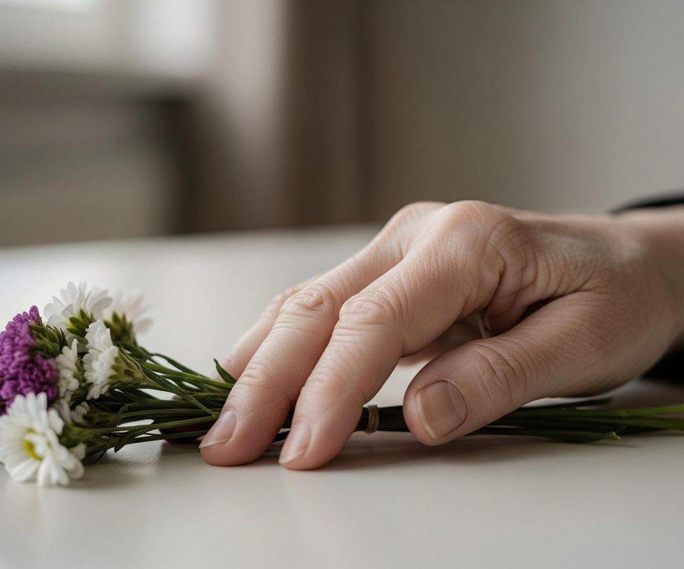 Dorosła dłoń z wyprostowanymi palcami leży na białym stole obok małego bukietu kwiatów. Dłoń ma jasną karnację i widoczne zmarszczki, z lekko zagiętym kciukiem i opuszkiem palca wskazującego dotykającym łodyg kwiatów. Bukiet składa się z białych i fioletowych kwiatów o drobnych płatkach, związanych jasnobrązową gumką wokół zielonych łodyg. Tło jest rozmyte, z niewyraźnymi odcieniami beżu i brązu, a z prawej strony widoczny jest fragment ciemnego materiału.