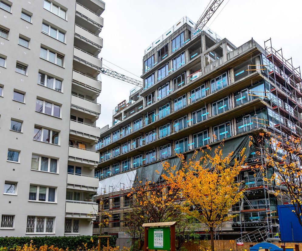 Wielorodzinny budynek w trakcie budowy przy ulicy Mordechaja Anielewicza w Warszawie, z widocznym żurawiem i rusztowaniami. Obok stoi starszy blok mieszkalny, a na pierwszym planie jesienne drzewa. Więcej o tej kontrowersyjnej inwestycji przeczytasz na Architektura Murator Plus.