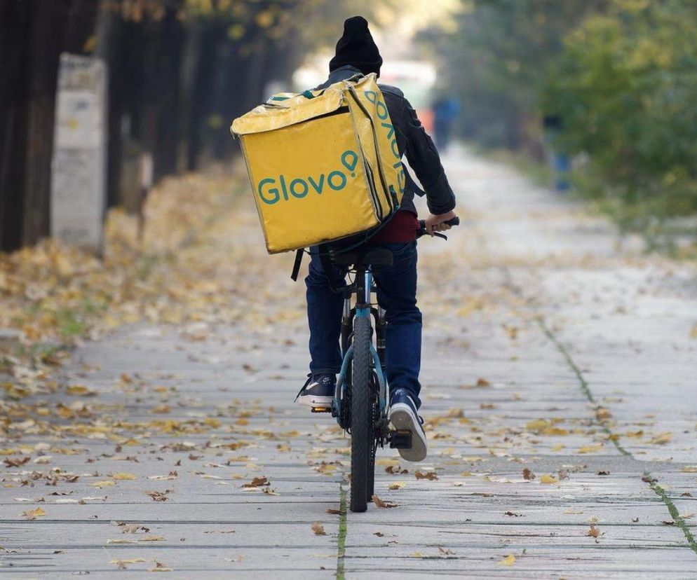 Kurier rowerowy Glovo z żółtym plecakiem dostarczający przesyłkę w otoczeniu jesiennych liści. Ilustracja symbolizuje rynek pracy cudzoziemców i jego wyzwania, o czym przeczytasz na portalu Super Biznes.