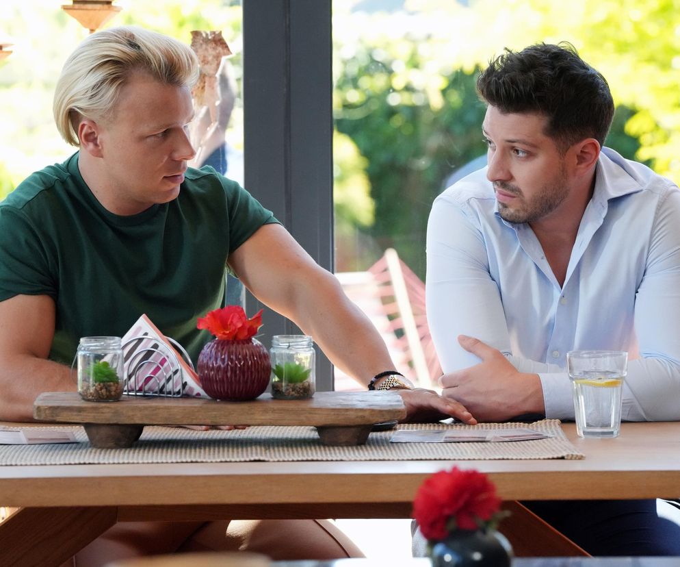 Patryk (Konrad Skolimowski) i Justin (Jasper Sołtysiewicz) z serialu Barwy szczęścia siedzą przy stole w kawiarni. Patryk, w zielonej koszulce, ma jasne włosy i patrzy w lewo, z lewą ręką opartą o stół, podczas gdy Justin w jasnoniebieskiej koszuli z ciemnymi włosami patrzy na niego z wyrazem zamyślenia. Na stole widoczne są ozdobne świeczki i szklanka z wodą, a tło stanowią rozmyte zielone liście drzew, co dodaje obrazowi naturalności. Zdjęcie to ilustruje fragment artykułu o serialu Barwy szczęścia dostępny na portalu Super Seriale.