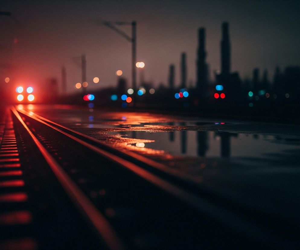 Torowisko biegnie przez środek kadru, z lewej strony widoczne są szyny oświetlone czerwonawym światłem. W oddali, na końcu torów, majaczy kształt pojazdu z czterema jasno świecącymi, białymi światłami w czerwonej poświacie. Prawa strona torowiska jest mokra, odbijając rozmazane, kolorowe światła z tła: pomarańczowe, czerwone i niebieskie. Na horyzoncie rysują się niewyraźne, ciemne sylwetki wysokich budynków przemysłowych, a nad nimi wiszą rozproszone światła latarni.