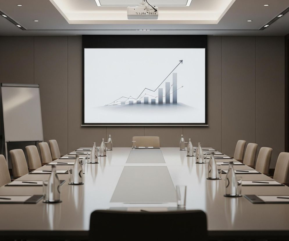 W centrum obrazu znajduje się długi stół konferencyjny w kształcie prostokąta, wykonany z jasnego, błyszczącego materiału. Po obu stronach stołu rozmieszczone są beżowe krzesła, a na blacie widoczne są szklane butelki z wodą, szklanki oraz notatniki z długopisami, ułożone symetrycznie. Na wprost stołu, na ścianie, wisi duży, jasny ekran projekcyjny z czarną ramą, na którym wyświetlany jest szary wykres słupkowy i liniowy ze strzałką skierowaną w górę. Nad ekranem, przymocowany do sufitu, widać rzutnik multimedialny. Po lewej stronie ekranu, blisko ściany, stoi biała, pionowa tablica flipchart. Prawa strona pomieszczenia jest zamknięta ciemnymi, drewnianymi drzwiami. Całe pomieszczenie jest utrzymane w neutralnych kolorach beżu i szarości, oświetlone punktowo z sufitu.
