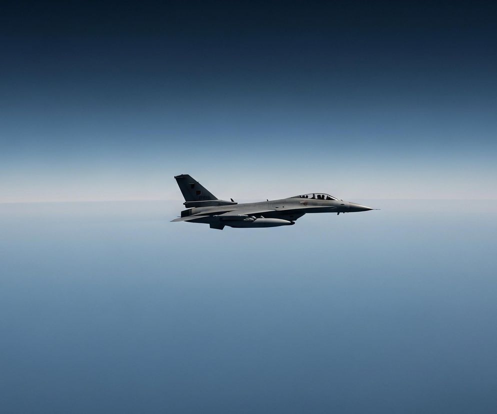 Szary samolot odrzutowy unosi się w powietrzu, lecąc w lewą stronę, równolegle do horyzontu. Posiada dwa podwieszane zbiorniki paliwa pod skrzydłami oraz pionowy stabilizator z tyłu. Tło stanowi błękitne niebo, przechodzące od ciemnego granatu u góry do jaśniejszego błękitu i bieli przy dolnej krawędzi obrazu, gdzie niewyraźnie widać linię horyzontu.