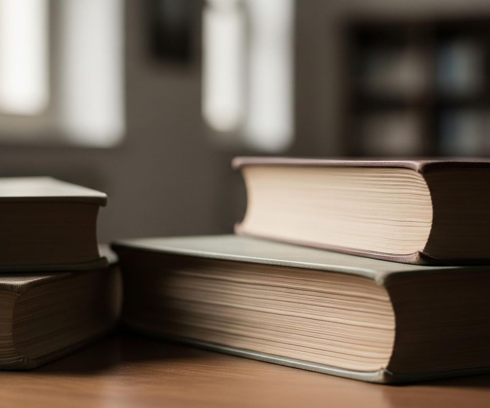 Na drewnianym stole leżą cztery książki o różnej grubości. Trzy książki są ułożone w stosy, dwie z nich po lewej stronie tworzą niski stos, a jedna książka leży na innej po prawej stronie. Okładki książek są w odcieniach szarości i brązu, a ich stronice mają jasny, kremowy kolor. Tło jest rozmyte, z widocznym jasnym prostokątem po lewej stronie, przypominającym okno lub oświetloną ścianę, oraz ciemniejszą, niewyraźną półką z książkami po prawej.