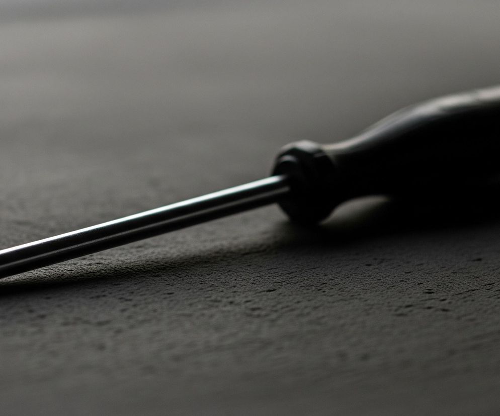 Śrubokręt z czarną, profilowaną rączką i srebrnym, metalowym trzonkiem leży pod lekkim kątem na ciemnej, szorstkiej powierzchni. Metalowa końcówka śrubokręta, typu krzyżak, jest skierowana w lewo, a trzonek zwęża się w kierunku rączki po prawej stronie. Na powierzchni widać delikatne cienie i fakturę, z jaśniejszym obszarem w górnej części tła.