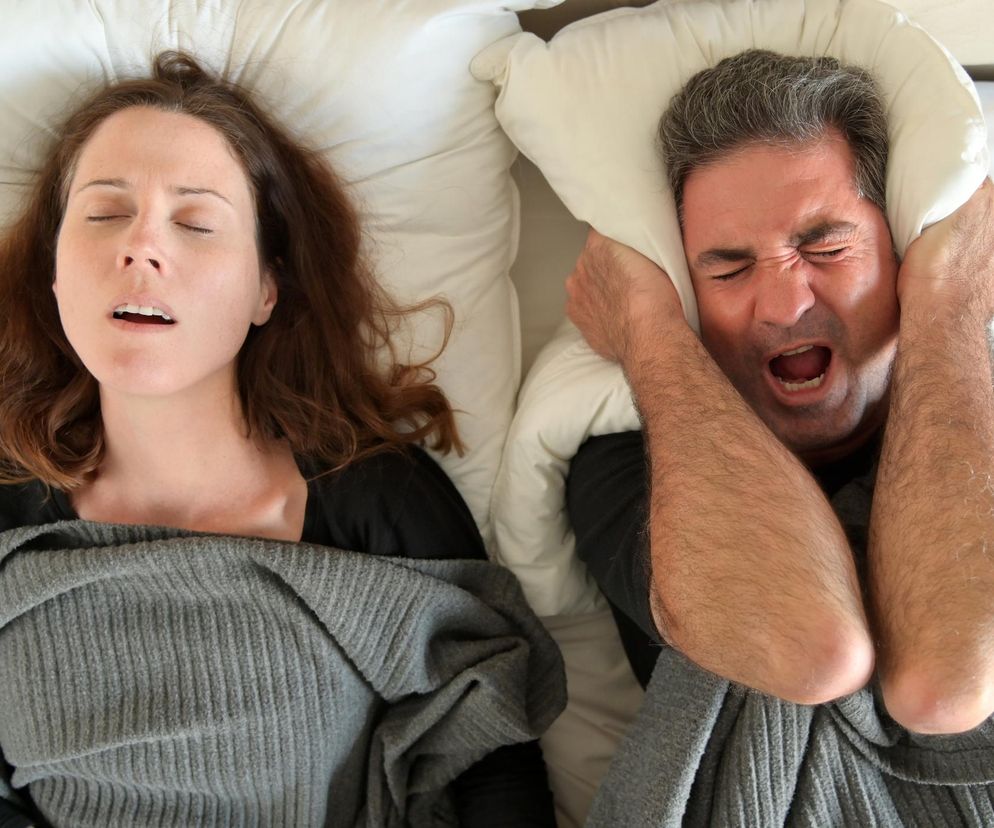 Kobieta chrapiąca z otwartymi ustami obok sfrustrowanego mężczyzny, który zasłania uszy poduszką. Scena symbolizuje problem chrapania w związku. Na temat chrapania, kto częściej chrapie – kobiety czy mężczyźni, oraz jak sobie z tym radzić, można przeczytać na portalu Poradnik Zdrowie.