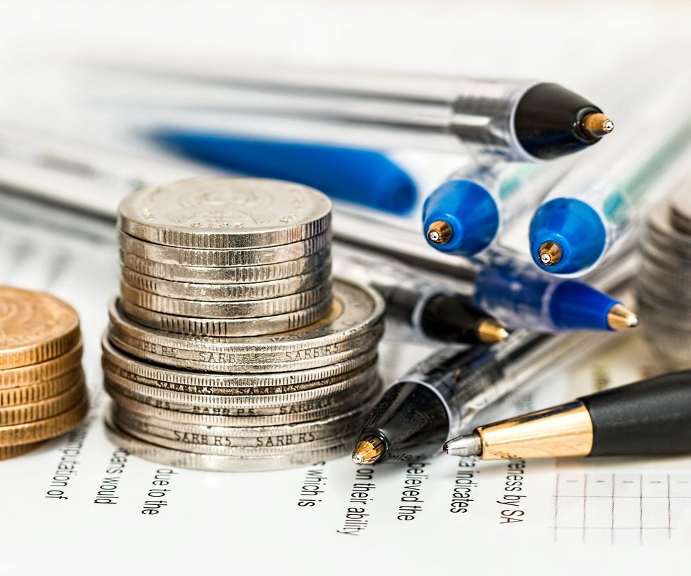 Stos monet ułożonych na dokumentach finansowych oraz długopisy. Symbolizuje niegospodarność i afery finansowe, jak ta opisana w Super Biznes o ubezpieczycielu Benefia, który stracił miliony na nieudanych inwestycjach. Zdjęcie ilustruje konsekwencje złych decyzji biznesowych.