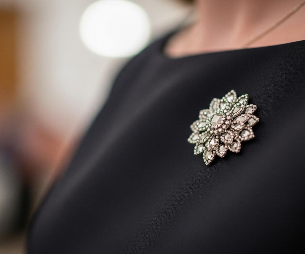 Na ciemnogranatowej tkaninie sukienki widoczna jest ozdobna broszka w kształcie stylizowanego kwiatu, bogato wysadzana licznymi, błyszczącymi kamieniami. Centralny element broszki stanowi większy, kwadratowy kamień otoczony mniejszymi, okrągłymi. Dalsze, podłużne kamienie ułożone są promieniście, tworząc efekt płatków. Górna część kadru ukazuje fragment obojczyka i dekoltu osoby ubranej w sukienkę, z delikatnym, złotym łańcuszkiem spoczywającym na skórze. Tło jest rozmyte, z jasnym, okrągłym punktem światła w górnej części.