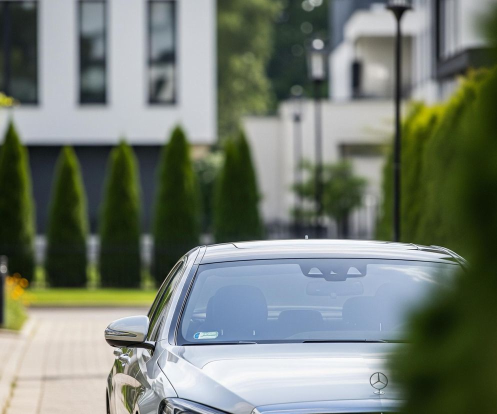 Srebrny samochód stoi zaparkowany na brukowanej ścieżce, z wyraźnym widokiem na jego przednią maskę, przednią szybę i lusterko boczne. Na masce widnieje logo Mercedes-Benz. Tło jest rozmyte, ukazując nowoczesne białe budynki z ciemnymi oknami oraz szereg zielonych stożkowatych krzewów. W prawym dolnym rogu oraz częściowo z lewej strony widoczne są rozmyte zielone liście, co sugeruje perspektywę spoza zarośli.