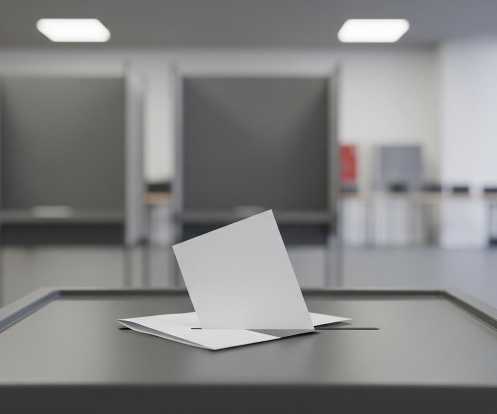 Na pierwszym planie, centralnie, widoczne jest górne wieczko ciemnoszarej urny wyborczej, z otwartą szczeliną, do której częściowo włożona jest biała koperta z kartą do głosowania. Koperta wystaje pionowo ze szczeliny, z widoczną przednią stroną, a jej dolna część jest zagięta pod kątem, spoczywając na wieczku urny. Po prawej stronie urny, na krawędzi wieczka, znajduje się cienki, czarny element w kształcie litery „L”. Tło jest rozmyte, ukazując szare kabiny do głosowania ustawione w rzędzie, a za nimi białe ściany i duże okna, wpuszczające dzienne światło. Sufit jest jasnoszary z prostokątnymi, białymi lampami.