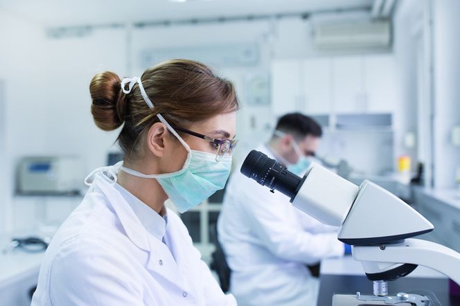 Laborantka w masce medycznej i okularach pochylona nad mikroskopem w sterylnym laboratorium. W tle, drugi naukowiec również pracuje przy sprzęcie laboratoryjnym. Scena symbolizuje intensywne badania nad szczepionkami, jak te opisywane na Poradnik Zdrowie.